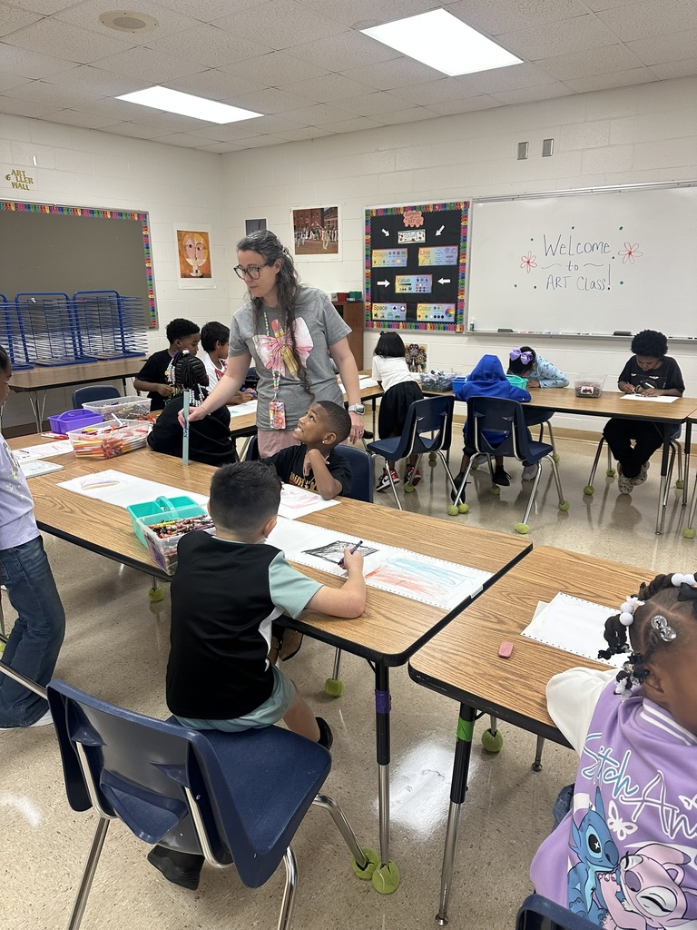 An art teacher helps a student at a table while classmates color and draw during art class.