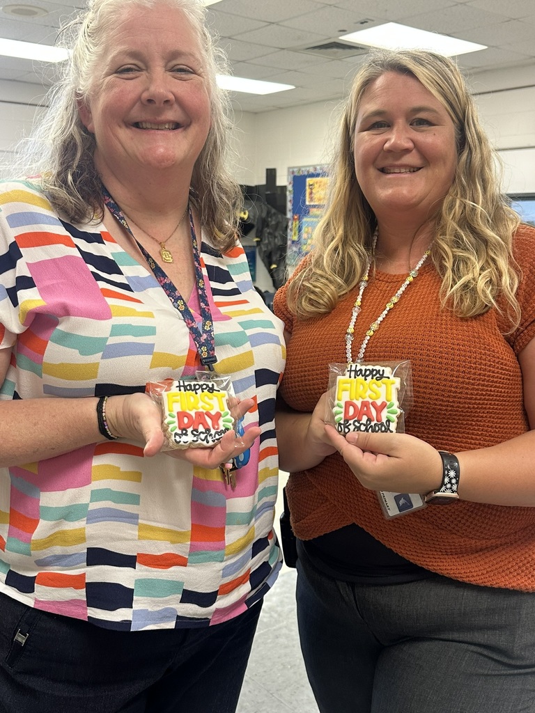 Two teachers hold colorful “Happy First Day of School” cookies and smile.