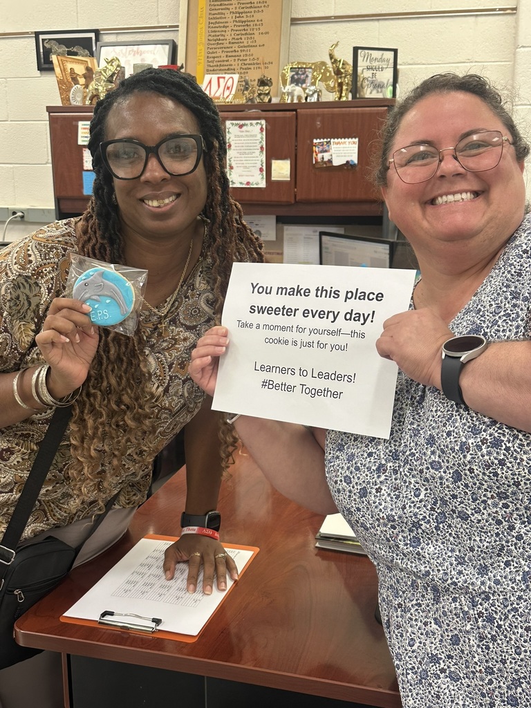 Two staff members smile, holding a dolphin cookie and a sign that reads, “You make this place sweeter every day!”