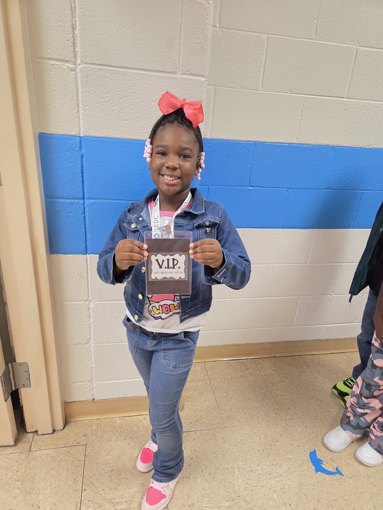 A smiling girl stands in a school hallway, proudly holding a laminated card labeled “V.I.P.” She wears a denim jacket and jeans, a white shirt with colorful designs, and bright pink sneakers with a large pink bow in her hair.