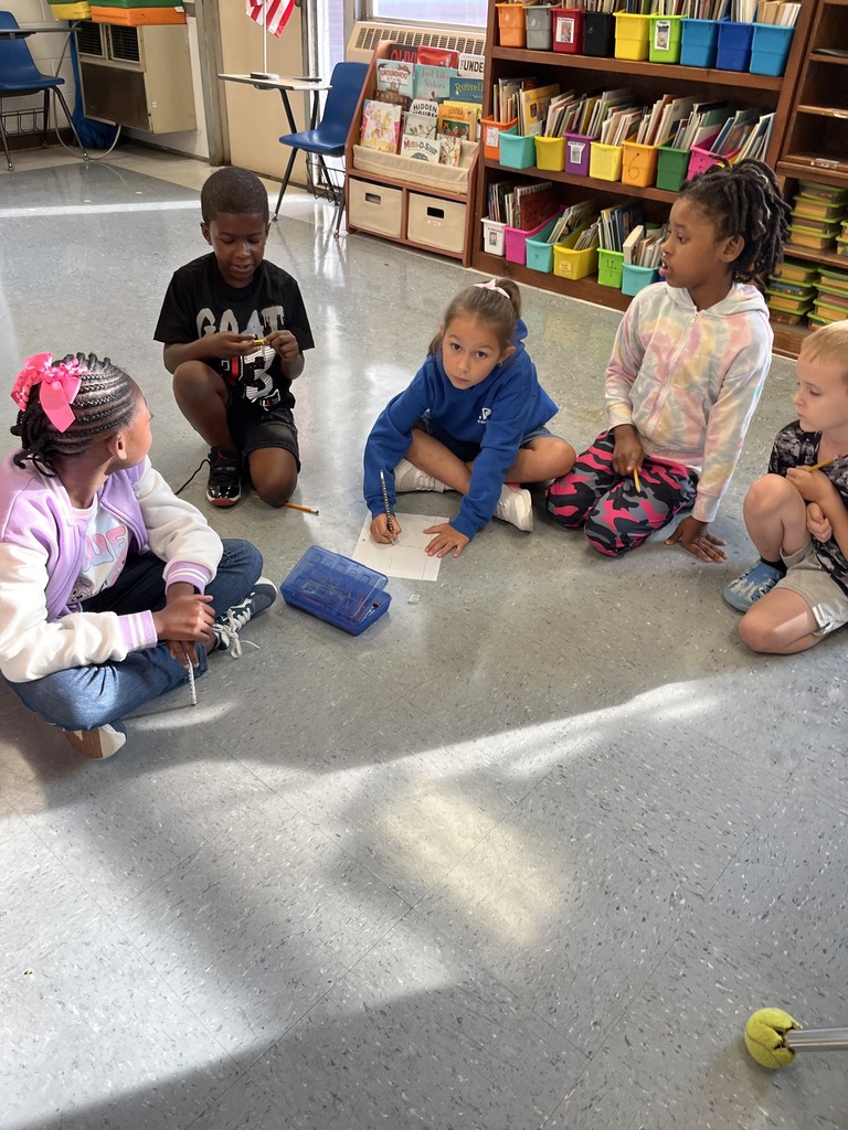 Five students sit together on the floor of a classroom, pencils in hand. They are smiling and focused as they brainstorm and write as a group, surrounded by colorful book bins