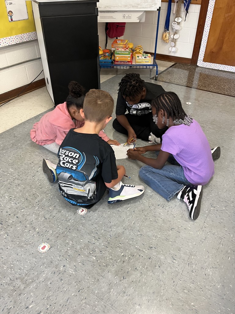 Four young students sit cross-legged in a circle on the classroom floor, leaning over a shared piece of paper as they work together. Two girls and two boys are focused, writing and discussing their ideas as a team.