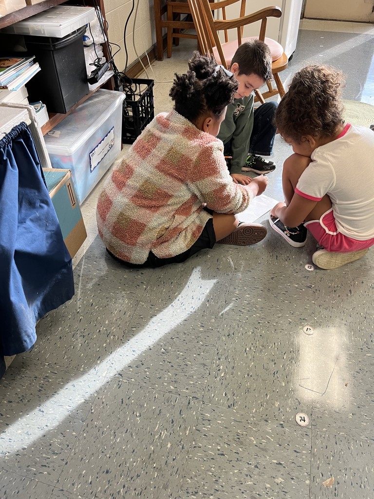 Three children crouch on the classroom floor in a small group. They lean in closely toward their work, collaborating on a paper activity near a wooden rocking chair and classroom storage area.