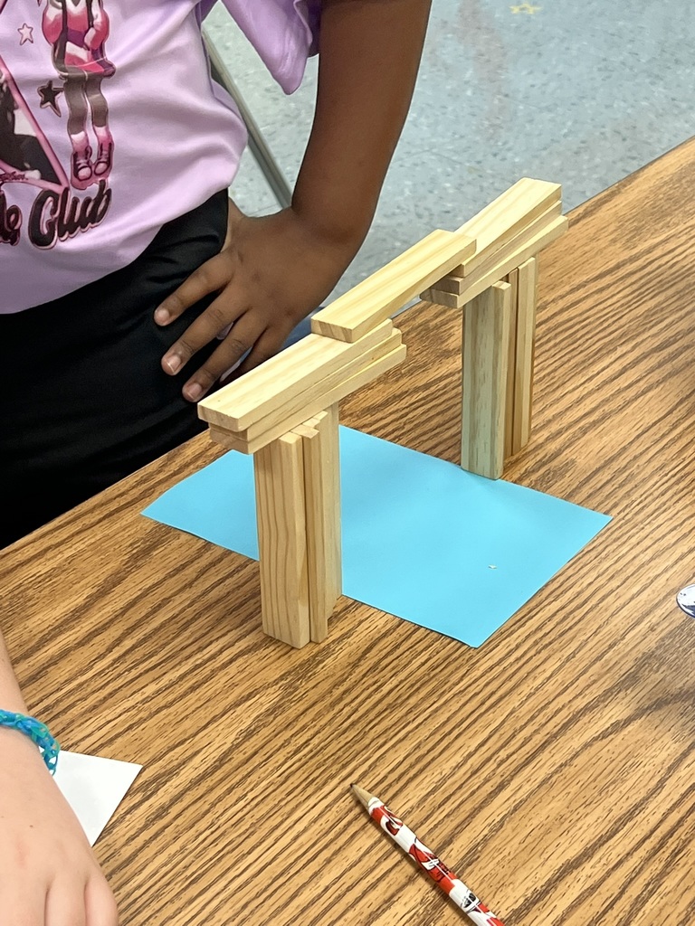 A finished student-built bridge made of wooden craft sticks stands proudly on a classroom table. The bridge, supported by two vertical pillars, spans across a bright blue piece of paper placed underneath. A student stands nearby, their hand resting on the table as they admire the structure. A pencil with a festive red-and-white design lies in front of the project.