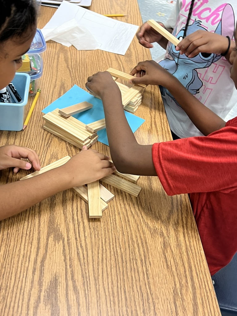 Several students sit around a classroom table, working together with wooden craft sticks. Their hands are busy stacking and arranging the sticks to form the base of a bridge structure. A blue piece of paper is placed under the project, and pencils, crayons, and supplies are scattered nearby.
