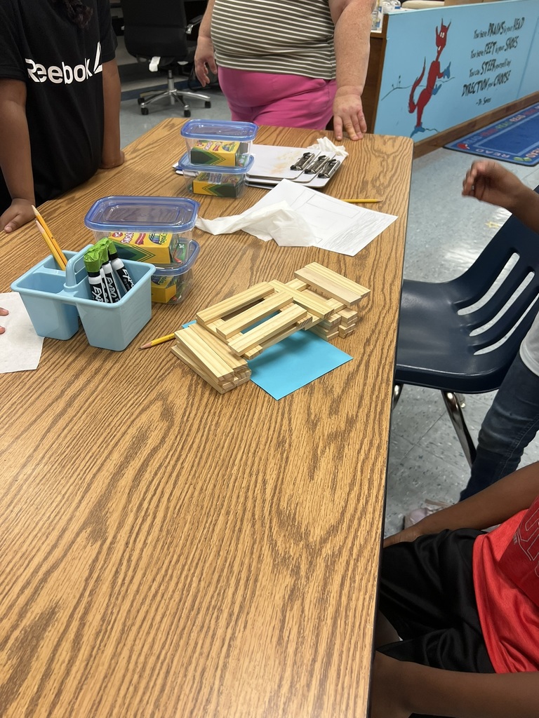 A group of students stand and sit around a wooden classroom table as they work on their bridge-building STEM challenge. On the table, a bridge made of wooden craft sticks rests on a bright blue piece of paper. The bridge is neatly stacked and shows structure forming in the middle.
