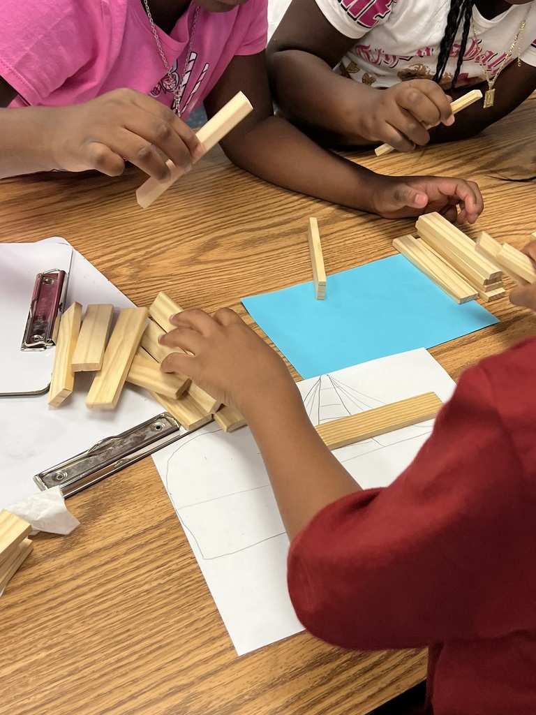 Close-up of students’ hands as they build a bridge with wooden craft sticks during STEM class. The sticks are being placed over a sheet of white paper with a bridge drawing sketched on it, showing planning before building. A blue paper square serves as the workspace background. Students in colorful shirts reach in from different sides of the table, carefully stacking and adjusting the sticks