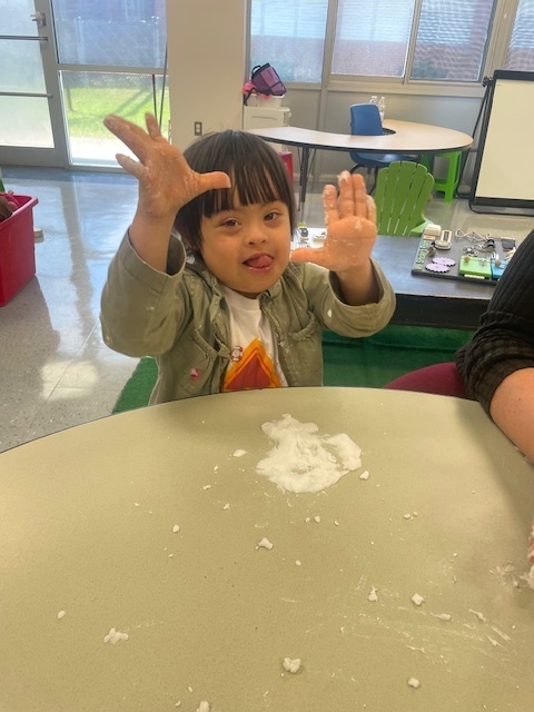 A young child sits at a table smiling with hands held up, covered in white sensory material, while playing during a classroom activity.