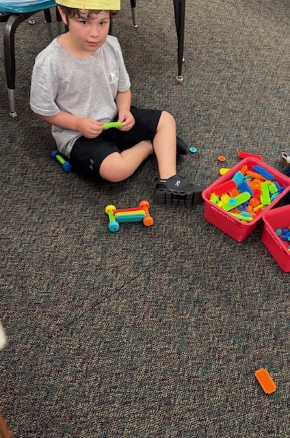 A young child wearing a gray shirt and black shorts is sitting on a carpeted classroom floor, playing with colorful plastic building toys. He is holding a green piece while several other pieces are scattered around him and in two red bins. A partially built structure is on the floor in front of him. The scene appears to be in a kindergarten or early childhood classroom.