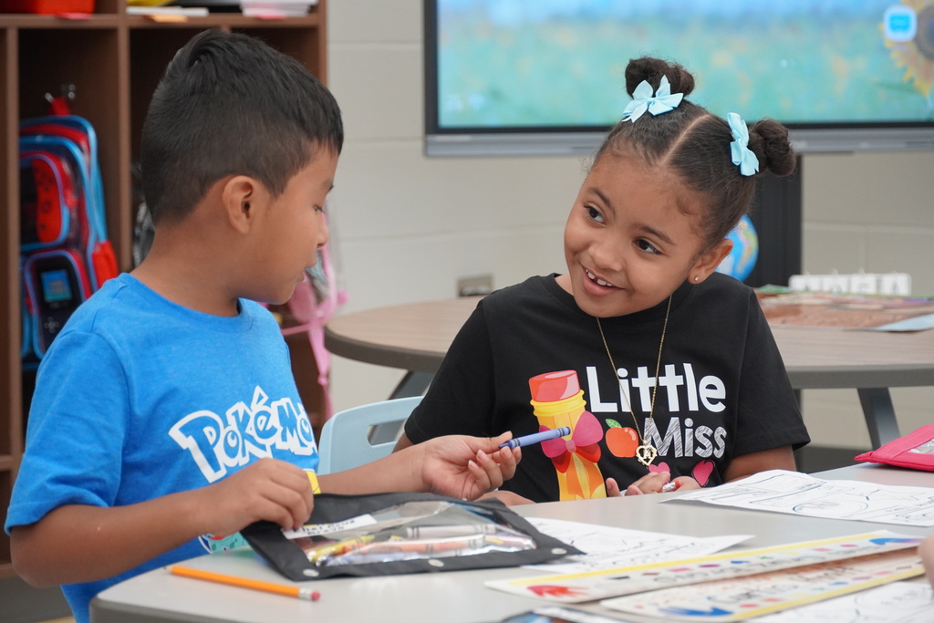 Two young students sit at a classroom table smiling and sharing markers while working on assignments