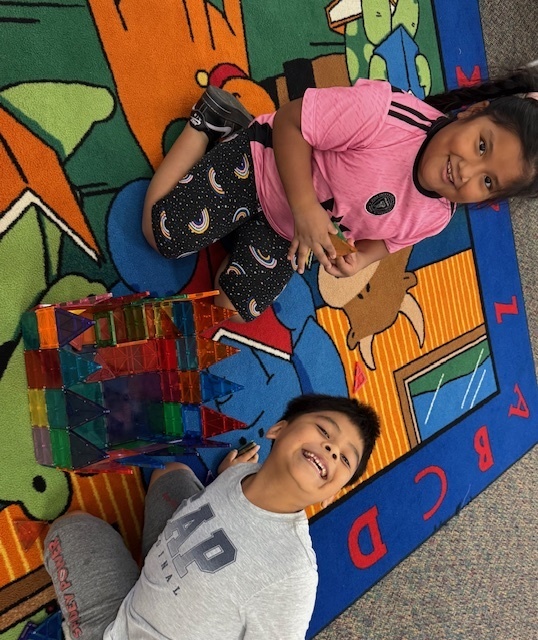 Two children are sitting and lying on a brightly colored alphabet rug, smiling at the camera. In front of them is a large, intricate structure made from colorful translucent magnetic tiles. The girl is wearing a pink shirt and patterned leggings, while the boy is in a gray GAP sweatshirt. The environment appears to be a classroom or library play area.