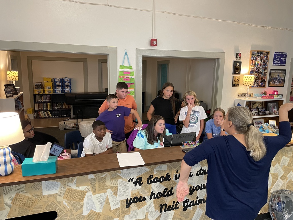 A group of middle school students gather around a library desk while a teacher speaks with animated gestures. The students appear engaged, some leaning forward or resting their chins in their hands. The desk is decorated with book pages and a quote from Neil Gaiman, and the background shows bookshelves, lamps, and classroom decor.