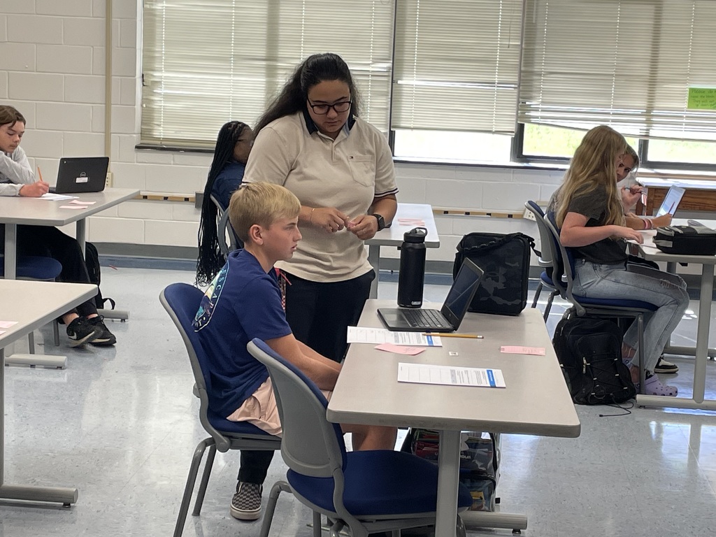 A teacher assists a student at his desk with a laptop open, while other students work independently in a classroom