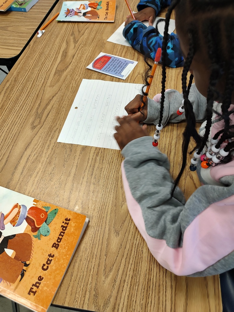 A student with braids and beads is writing on lined notebook paper with a pencil. Beside the paper is a card with a mailbox graphic and the prompt, “Write a letter to the principal telling how you would change the school.” A book titled The Cat Bandit is also on the desk.
