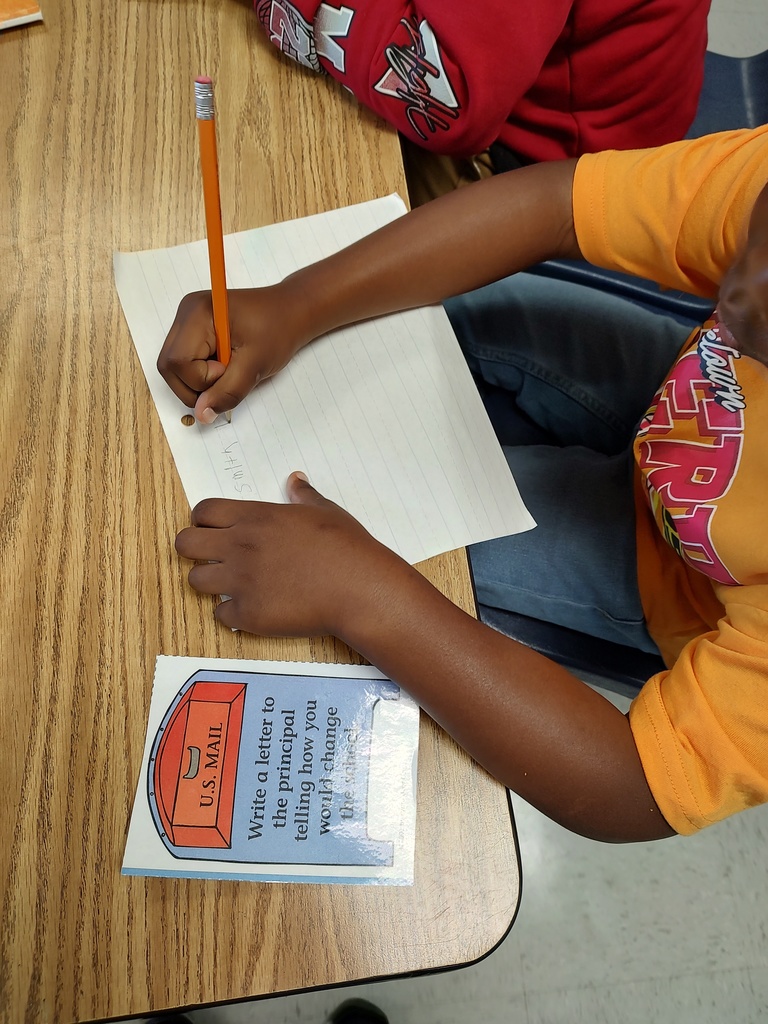A student in an orange shirt is writing on lined notebook paper with a pencil. On the desk is a card with a mailbox graphic that says, “Write a letter to the principal telling how you would change the school.”