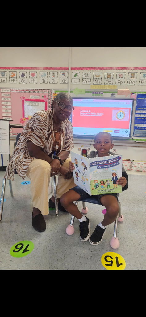 A kindergarten student sits proudly holding the book Superheroes Are Everywhere while reading aloud to the class. A smiling adult sits beside him, showing support and encouragement. The colorful classroom background includes alphabet cards, bulletin boards, and a digital screen with a lesson displayed. The moment highlights leadership, confidence, and a love of reading.