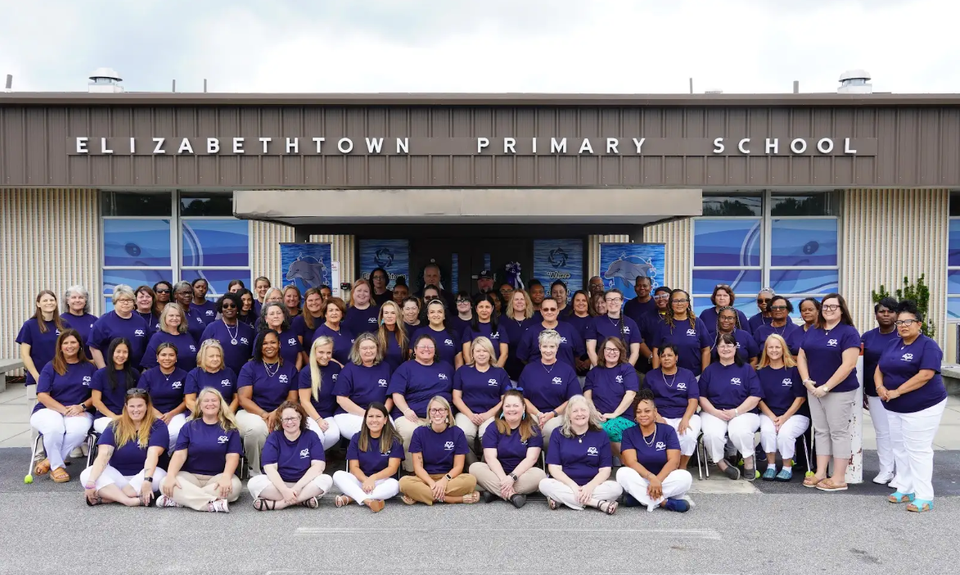A large group of staff members from Elizabethtown Primary School gather outside the school building for a group photo. Everyone is dressed in matching purple shirts and light-colored pants, smiling together to show unity and school spirit.