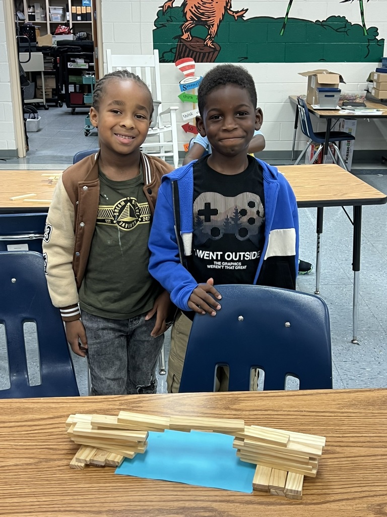 Two smiling students stand proudly behind their completed block bridge, which stretches across a piece of blue paper representing water. Their teamwork and creativity are on display.