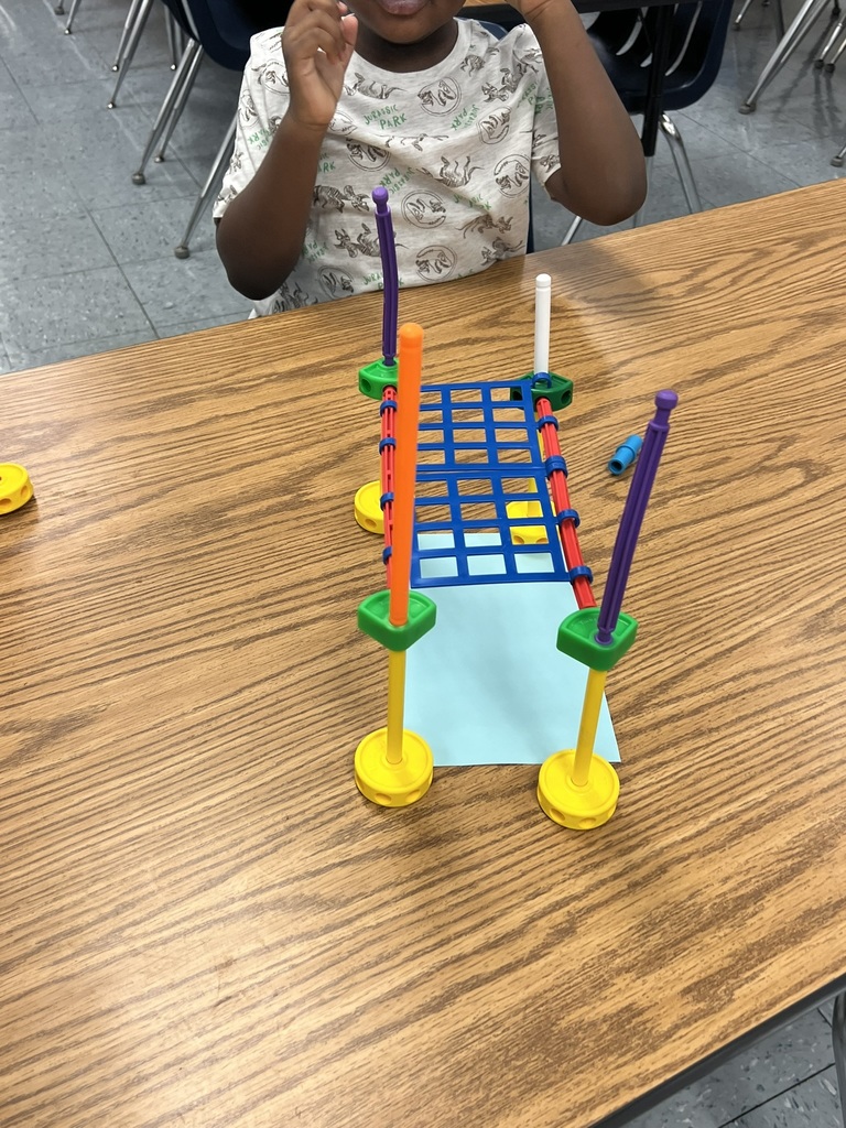 A student shows off a colorful bridge built with plastic construction pieces. The bridge features tall supports on each side and a grid-like platform spanning across a piece of blue paper.