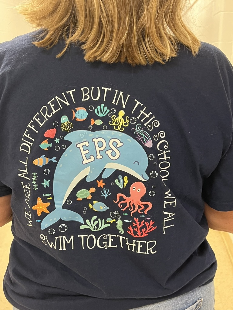 A person wearing a navy blue T-shirt with a colorful ocean-themed design on the back. At the center is a large light blue dolphin with “EPS” written across its body. Surrounding the dolphin are playful sea creatures, including an octopus, fish, turtle, seahorse, jellyfish, starfish, crab, and coral, along with seashells and bubbles. Above and around the dolphin, the text reads: “We are all different but in this school we all swim together.” The design emphasizes unity and diversity, using bright, cheerful sea life illustrations to create a friendly and welcoming message.