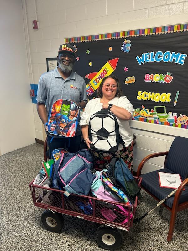 Two smiling adults stand in front of a colorful “Welcome Back to School” bulletin board, holding backpacks. The man wears a gray polo shirt and cap, holding a superhero-themed backpack. The woman, wearing glasses and a white shirt, holds a black-and-white soccer ball backpack. In front of them, a red wagon is filled with additional backpacks and school supplies, ready to be shared with students. The cheerful scene highlights their generous donation and community support for the school year.
