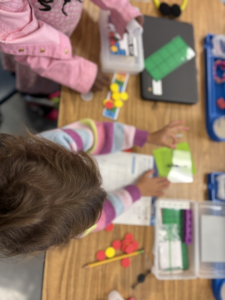 Two young students work together with math tool kits, using counters, ten-frames, and worksheets to solve equations.
