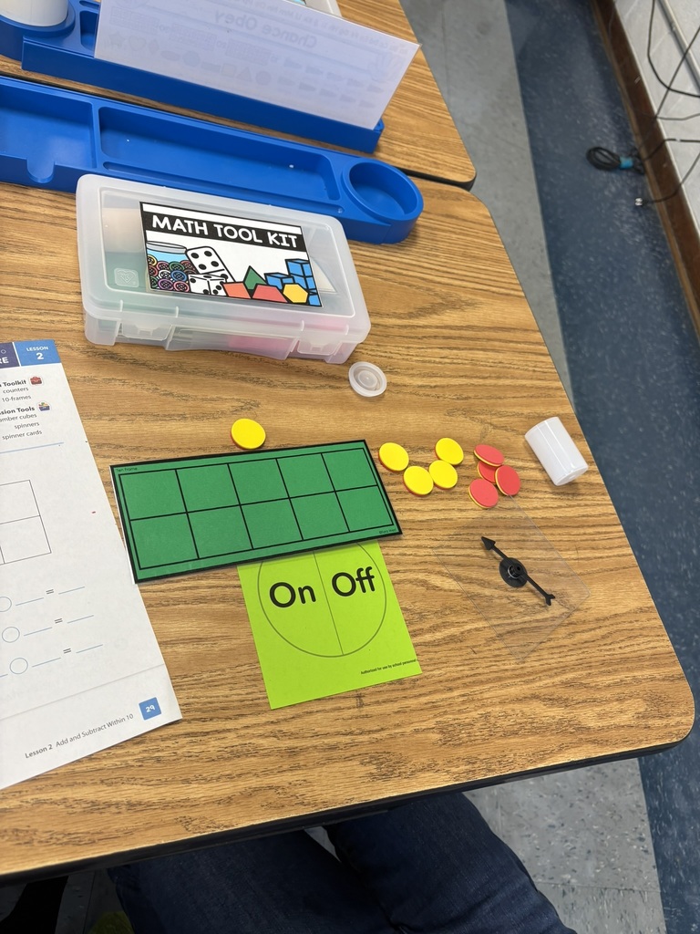 A classroom desk with a math tool kit box, red and yellow counters, a green ten-frame mat, and a worksheet for addition and subtraction.