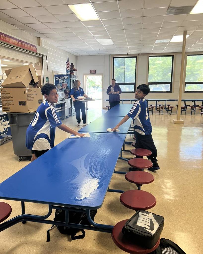 Two students wearing blue soccer jerseys wipe down a long cafeteria table with cloths, while two adults stand in the background near the serving line. The cafeteria has large windows letting in natural light and red round stools attached to the table.