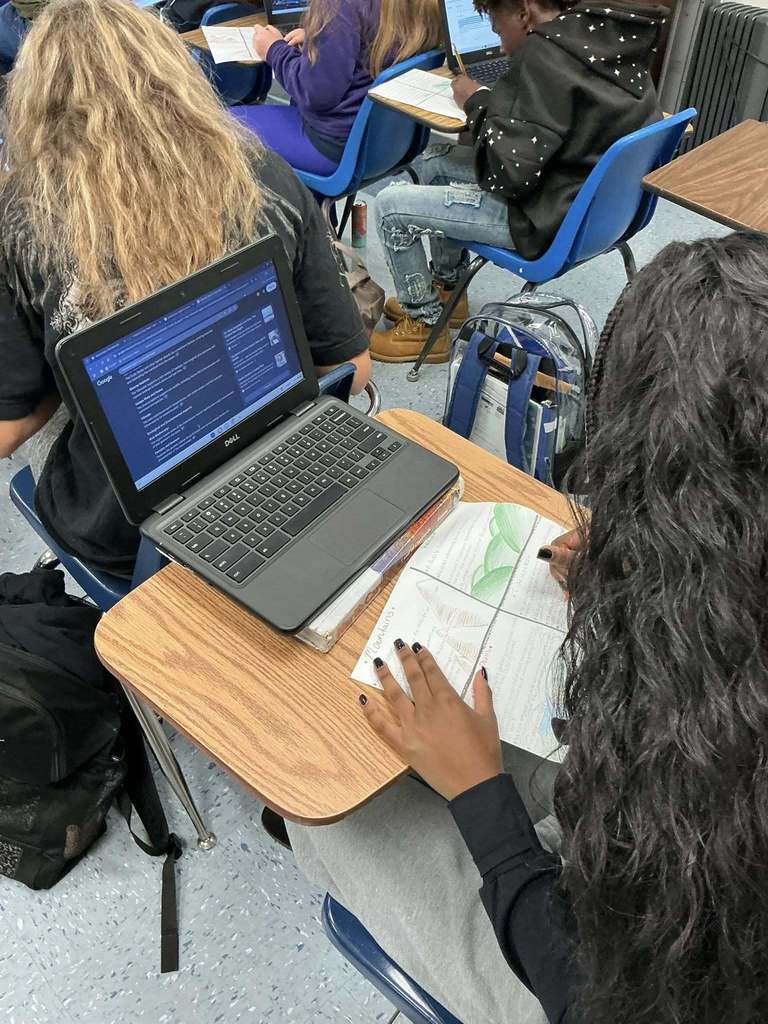 A student sits at a desk using both a Dell laptop and a notebook, working on an assignment that includes handwritten notes and drawings. Other students around the classroom are also writing in notebooks or using laptops.