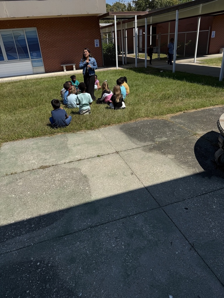 A teacher stands outside on the grass with a small group of elementary students sitting in a circle around her. The children are attentively listening as she leads a math lesson. The activity focuses on learning addition and subtraction within 10, taking advantage of the sunny day for outdoor learning. The school building and covered walkway are visible in the background, while the group enjoys hands-on instruction in a relaxed, open-air setting.