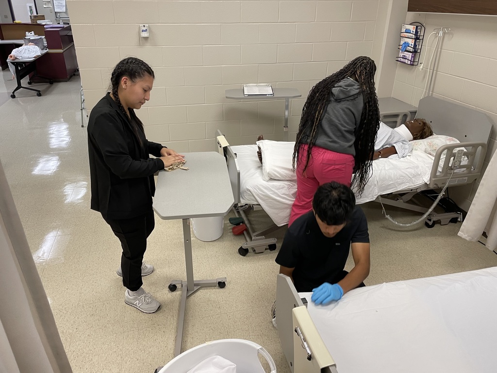 Three students practice healthcare skills in a classroom lab. One student folds supplies on a rolling table, another adjusts bedding for a mannequin patient lying in a hospital bed, and a third kneels at the foot of the bed wearing gloves while preparing the linens.