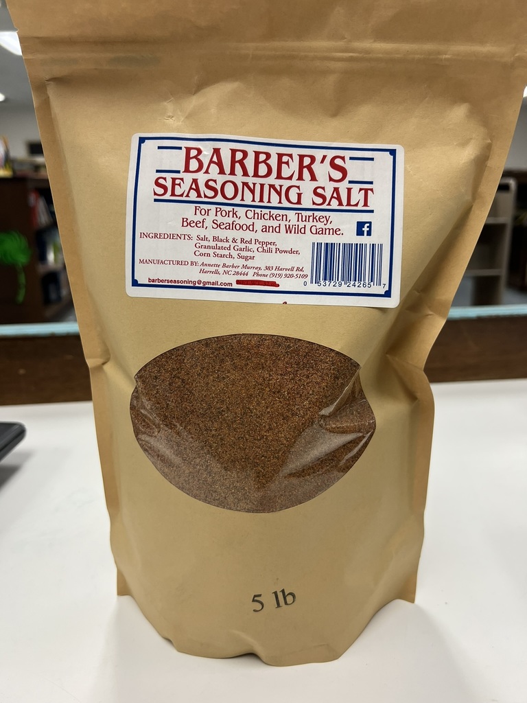 A 5-pound brown paper bag of Barber’s Seasoning Salt is displayed on a table. The label reads “Barber’s Seasoning Salt” in red and blue lettering and lists it as suitable for pork, chicken, turkey, beef, seafood, and wild game. The ingredients include salt, black and red pepper, garlic, chili powder, cornstarch, and sugar. A clear window on the bag shows the reddish-brown seasoning inside.