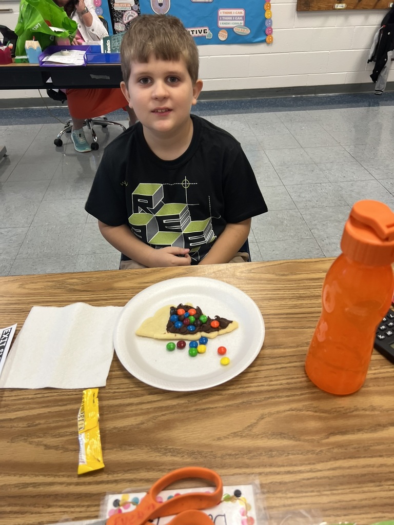 A young student sits at a desk smiling, with a North Carolina–shaped sugar cookie on a plate in front of him. The cookie is decorated with chocolate frosting and colorful M&Ms to represent the three regions of the state.