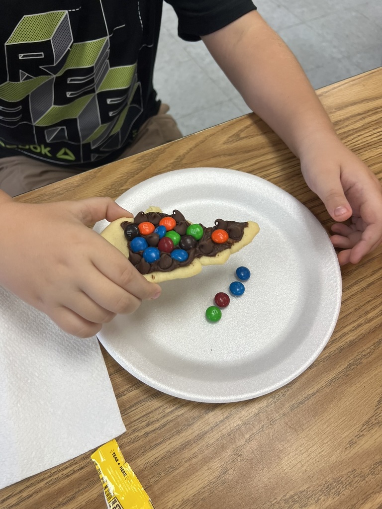 A close-up of a student’s hand holding a decorated cookie shows colorful M&Ms and chocolate chips spread across the state-shaped cookie to represent the three regions of North Carolina.