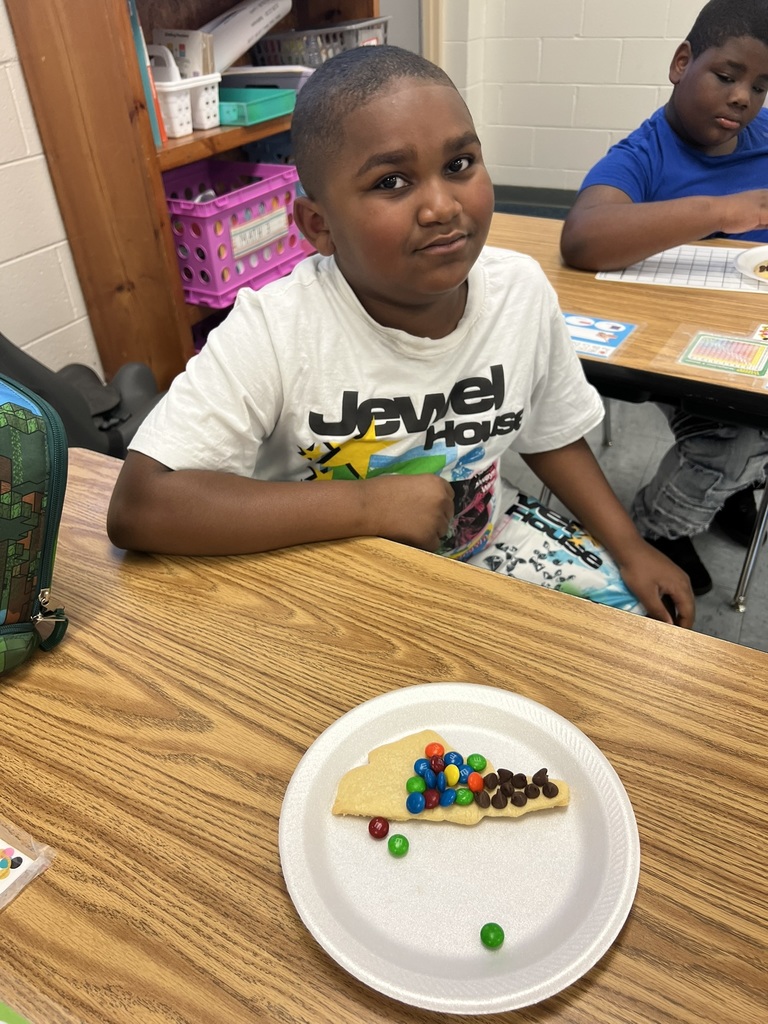 A student in a white shirt sits at his desk, looking at his decorated cookie. The cookie is covered with clusters of M&Ms and chocolate chips placed to represent North Carolina’s three regions.