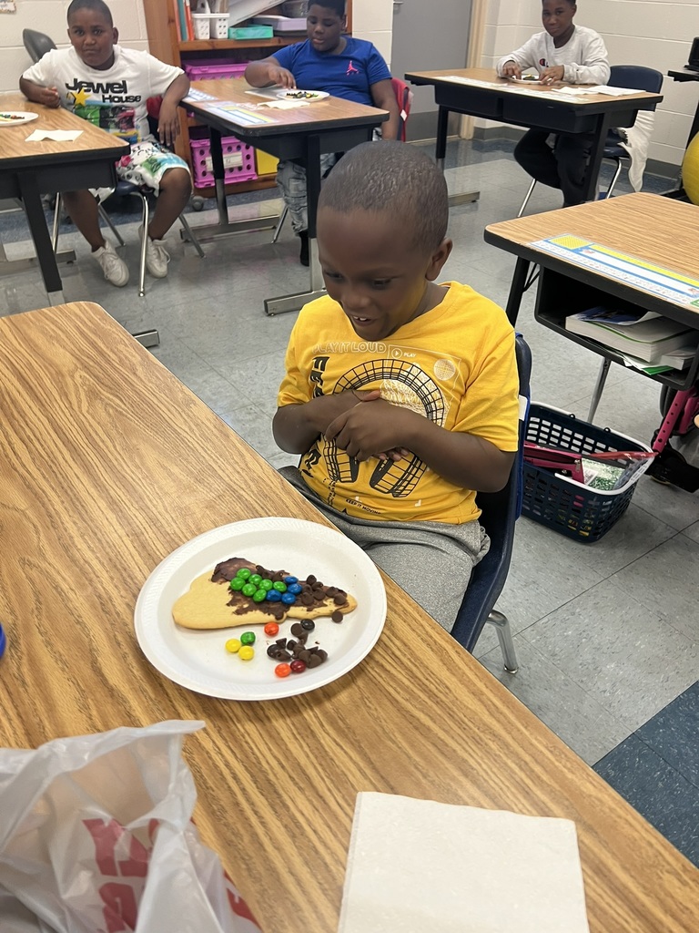 A student in a yellow shirt looks excited while sitting at a desk. In front of him is a cookie shaped like North Carolina, decorated with chocolate chips and M&Ms to show the three regions.