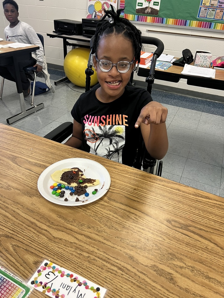 A smiling student wearing glasses and a black “Sunshine” shirt points proudly at her decorated cookie. Her cookie is spread with chocolate and candy pieces to illustrate North Carolina’s regions.