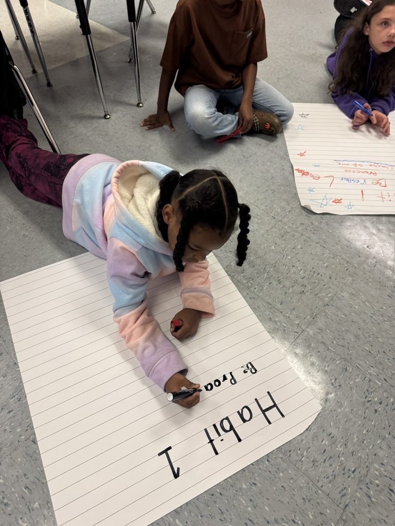 A student in a pastel hoodie writes “Habit 1” on a large chart paper while others sit nearby.
