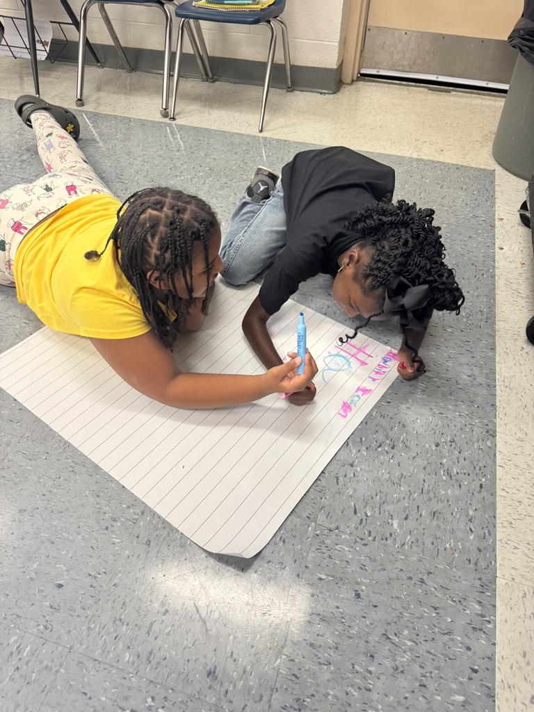 Two students lay on the floor working together with markers on a large sheet of paper, smiling and writing colorful words.
