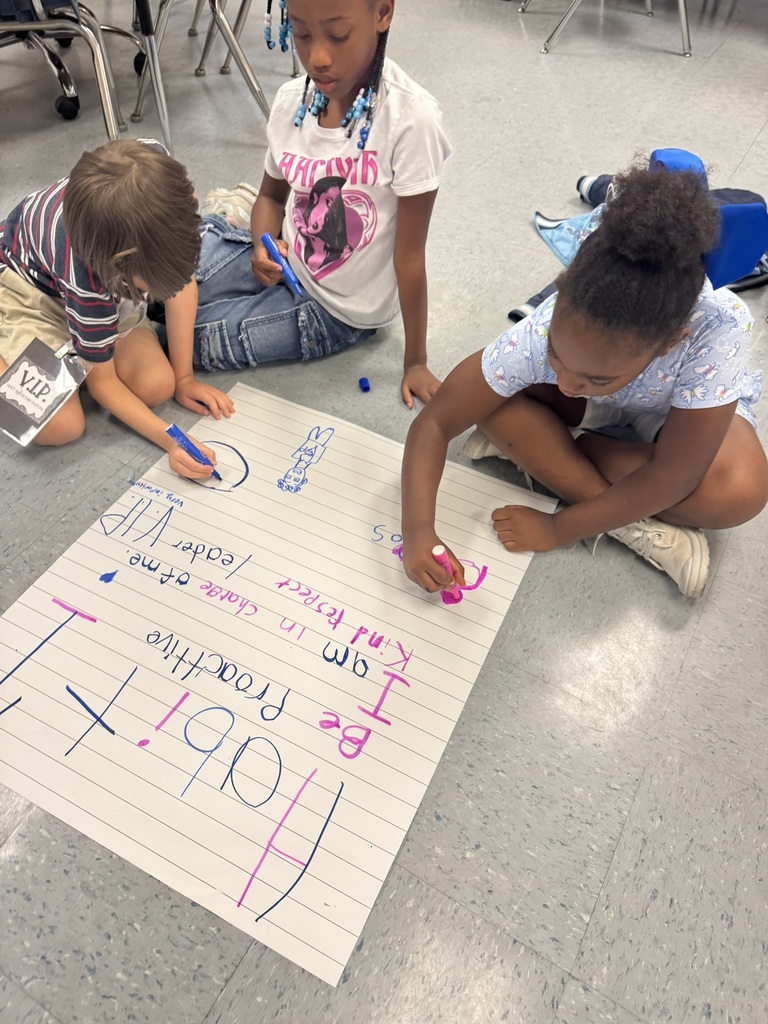 Three students sit in a group, writing and drawing on a poster about “Habit 1” with bright markers.