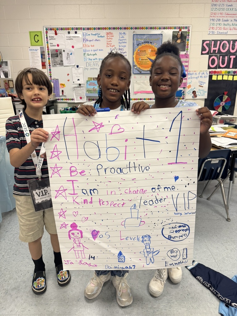 Three smiling students proudly hold up their finished Habit 1 poster decorated with words, stars, and drawings.