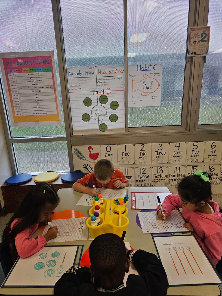 A small group of kindergarten students sit at a table near a window, practicing writing lines and shapes on whiteboards and worksheets.