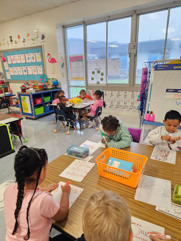 A larger group of kindergarten students works in centers, some tracing letters and shapes while others practice writing lines on their papers.