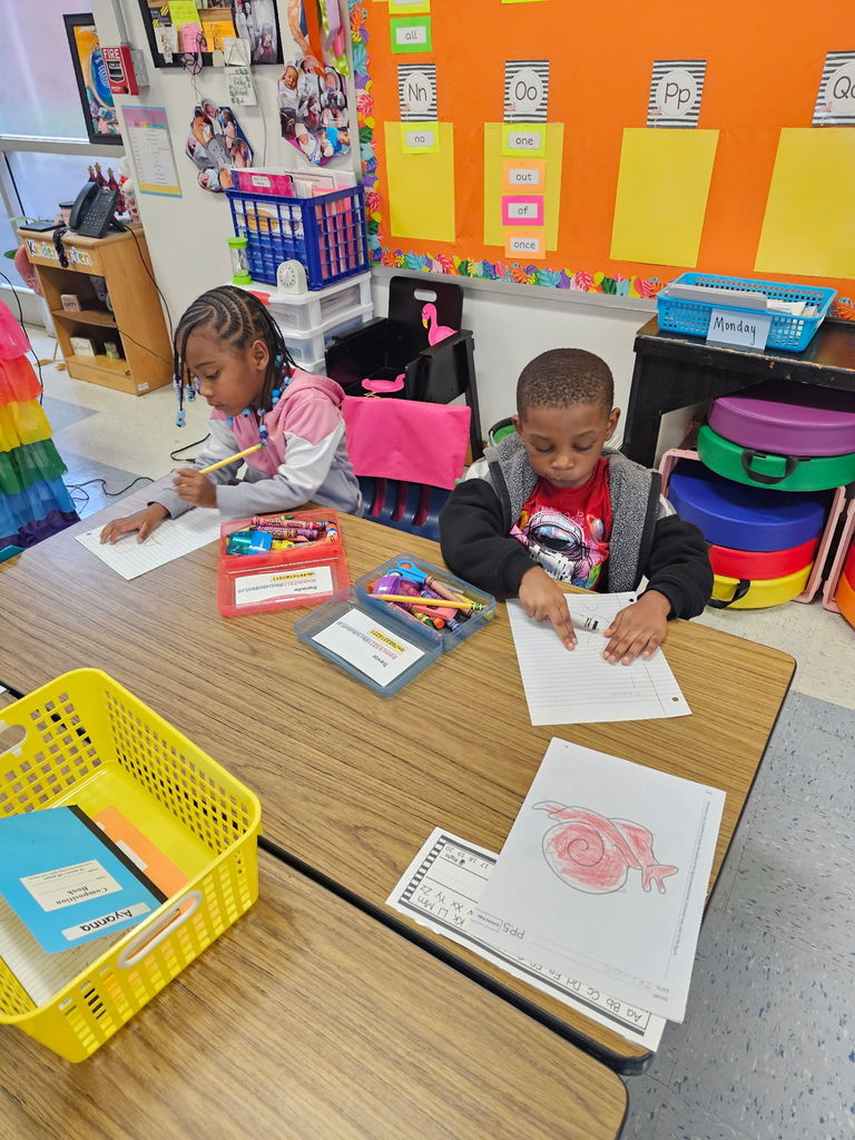 Two kindergarten students sit side by side at a table writing in their notebooks with crayons and pencils.