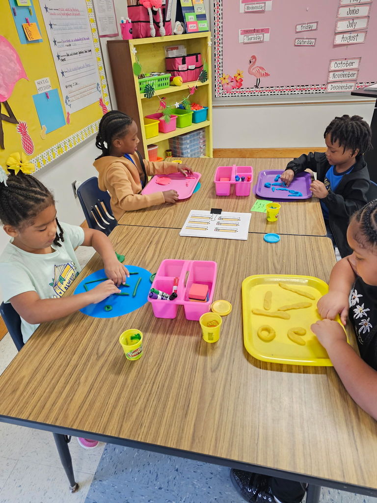 Four kindergarten students sit at a table using trays of Play-Doh to roll and shape lines, circles, and other patterns.