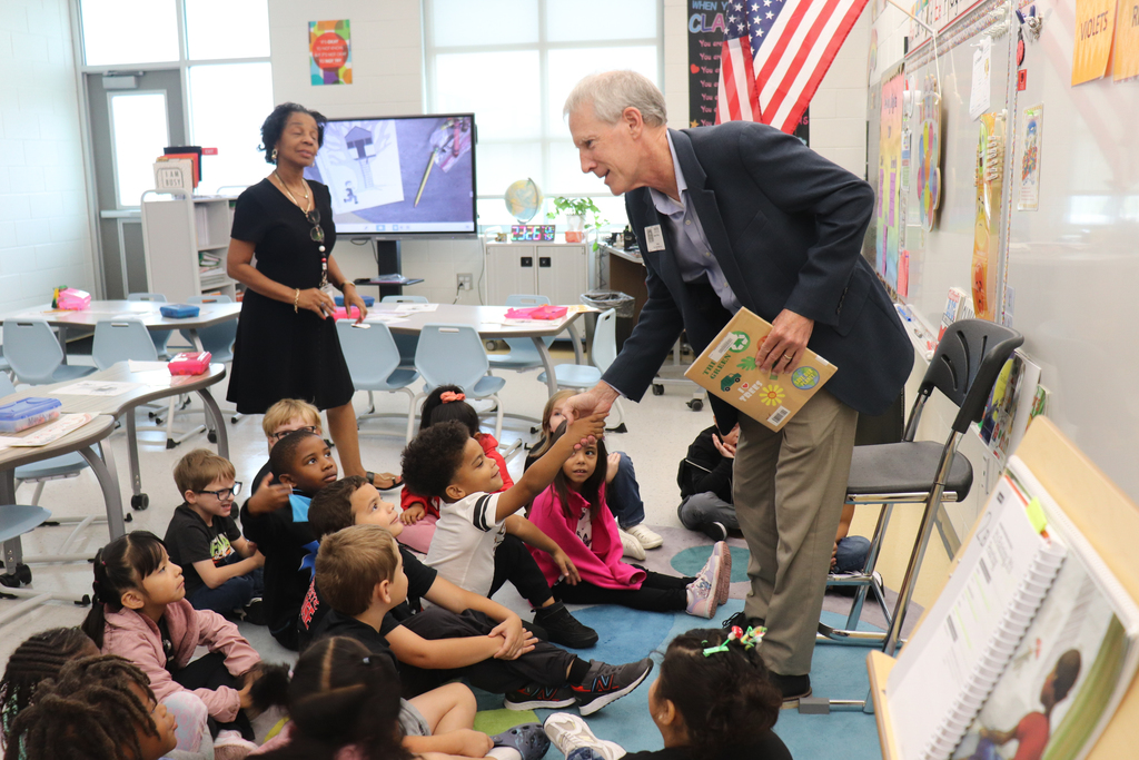 A man shakes hands with a young student seated on the floor of a classroom while holding a children’s book. Other students and a teacher look on with smiles.