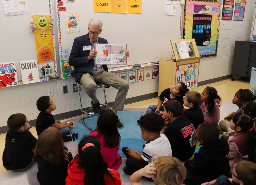 A man in a blazer speaks to a classroom of middle school students seated at desks with laptops. An American flag and digital screen are visible in the background.