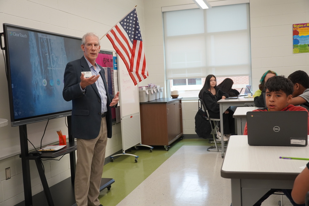 A man in a blazer speaks to a classroom of middle school students seated at desks with laptops. An American flag and digital screen are visible in the background.