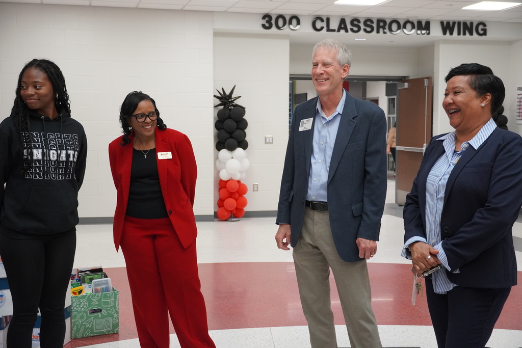A group of four people stand smiling together in a school hallway near a sign labeled "300 Classroom Wing." Black, red, and white balloon decorations are visible in the background.