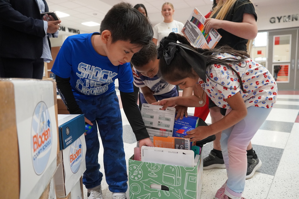 Three young students bend over to look through a supply box filled with papers, notebooks, and classroom items. Other children and adults stand nearby